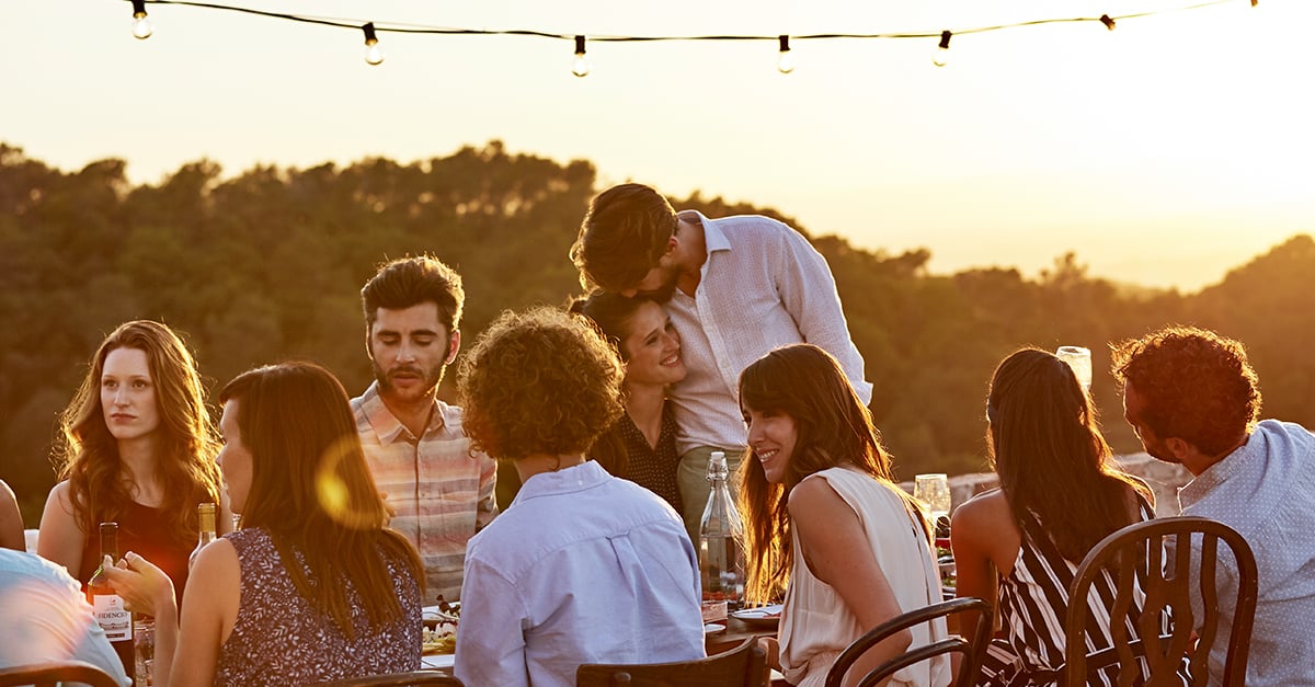 Gruppe von Menschen sitzt bei Sonnenuntergang zusammen und unterhält sich, ein Paar umarmt sich