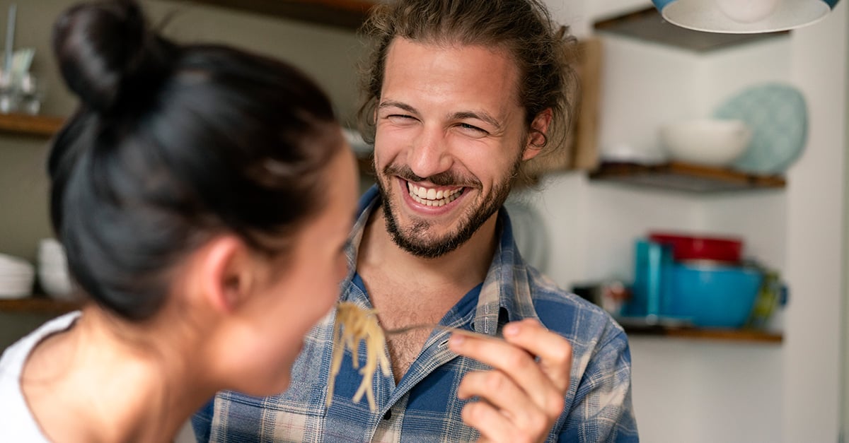 Ein Mann bietet einer Frau lachend eine Gabel mit Spaghetti an. Im Hintergrund ist eine Küche zu sehen.