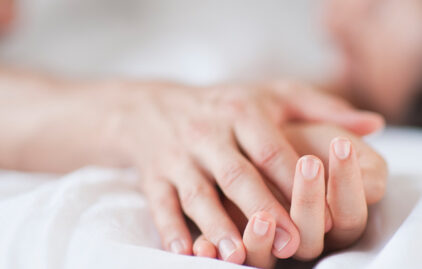Man sieht die Hände einer Frau und eines Mannes, die zärtlich verschränkt sind. Die Hände liegen auf einem weißen Bettlaken, das Bild hat eine erotische Atmosphäre.