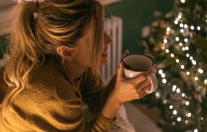 Eine junge Frau sitzt vor einem geschmückten Weihnachtsbaum, hält eine Tasse Tee in den Händen und lächelt. Das Bild vermittelt eine sehr gemütliche Atmosphäre.