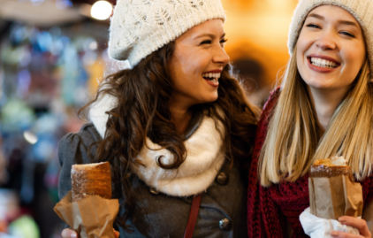 Zwei junge Frauen essen auf einem Weihnachtsmarkt Baumbrot. Sie lachen, fröhlich, tragen warme Mützen und dicke Schals.