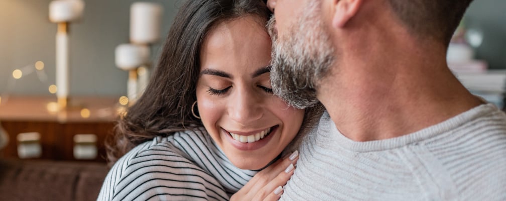 Eine Frau mit schwarzem Haar lehnt sich lächeln an die Schulter eines Mannes mit grau meliertem Bart