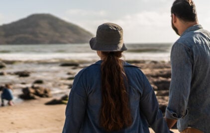 Man und Frau mittleren Alters stehen am Strand, halten sich an den Händen und beobachten zwei kleine Kinder, die im Sand spielen