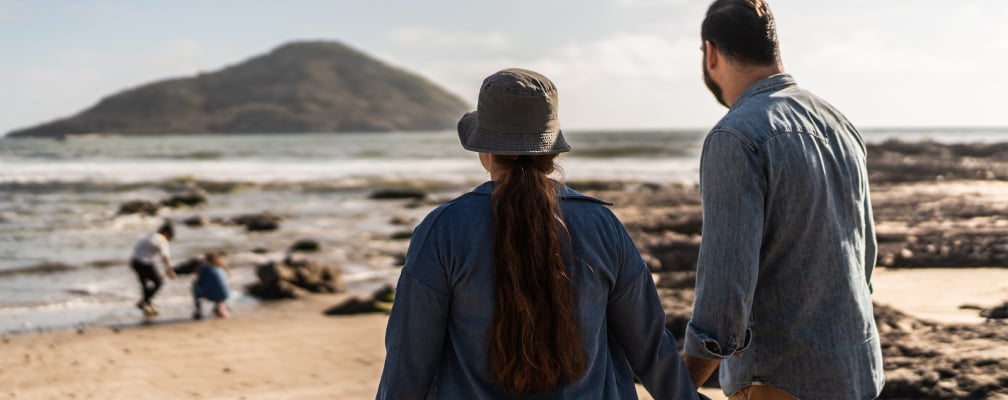 Man und Frau mittleren Alters stehen am Strand, halten sich an den Händen und beobachten zwei kleine Kinder, die im Sand spielen