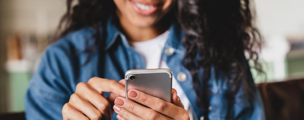 Im Vordergrund wird ein Handy gehalten, im Hintergrund ist eine lächelnde Frau zu erkennen.