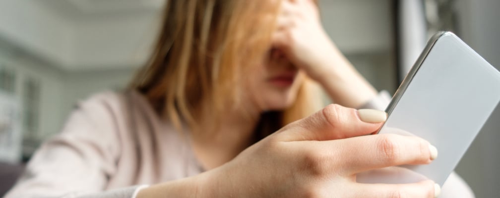 Im Vordergrund ist ein Smartphone zu sehen, im Hintergrund hält eine Frau frustriert ihren Kopf.