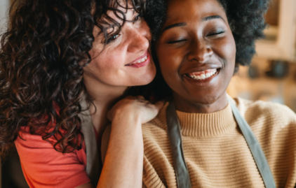 Eine weiße Frau mit Locken umarmt eine schwarze Frau in einem hellbraunen Pullover. Beide lächeln glücklich.