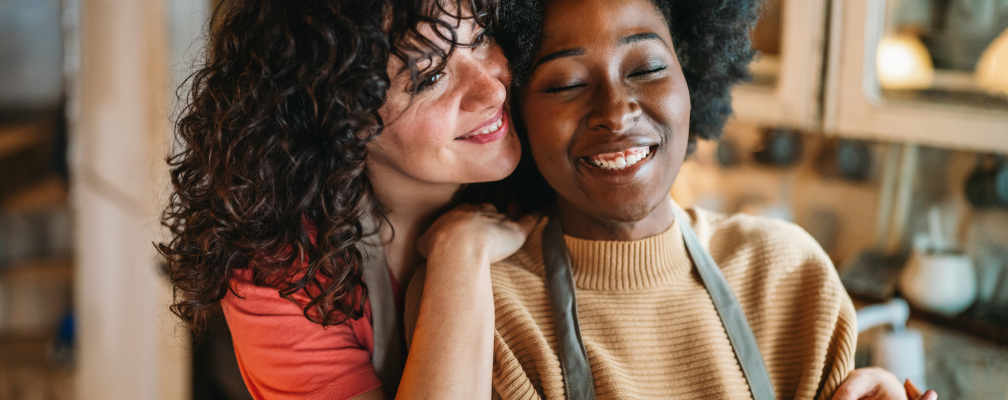 Eine weiße Frau mit Locken umarmt eine schwarze Frau in einem hellbraunen Pullover. Beide lächeln glücklich.